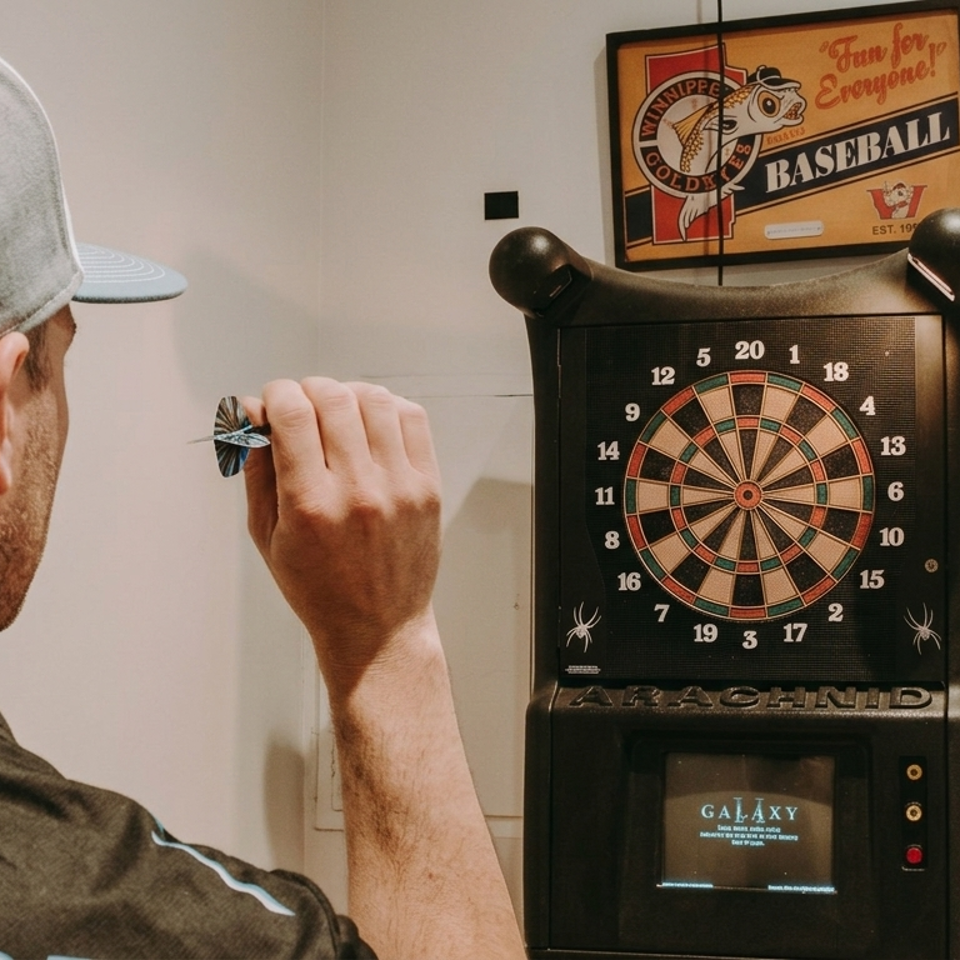 Dart player in a Winnipeg Dart League shirt aims at an electronic dartboard beneath a baseball sign.