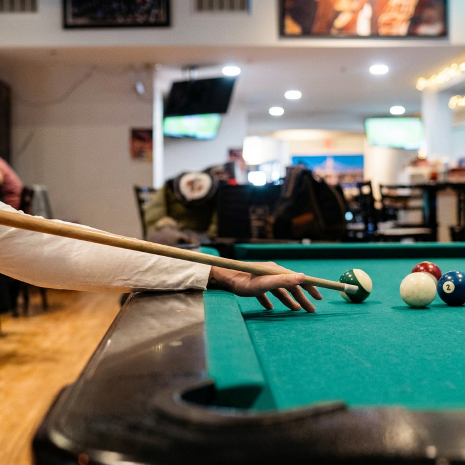 Person aiming a cue at billiard balls on a pool table inside a lively restaurant and bar with televisions.